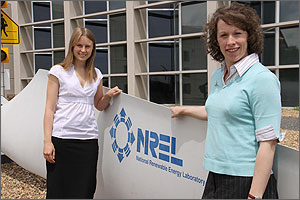 Photo of two women standing next to the white blade of a wind turbine, smiling at the camera.