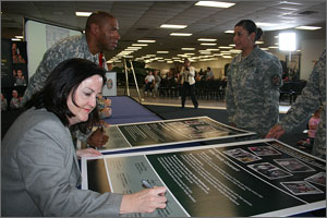 Photo of a woman signing a large poster with men and women in uniform in the background.