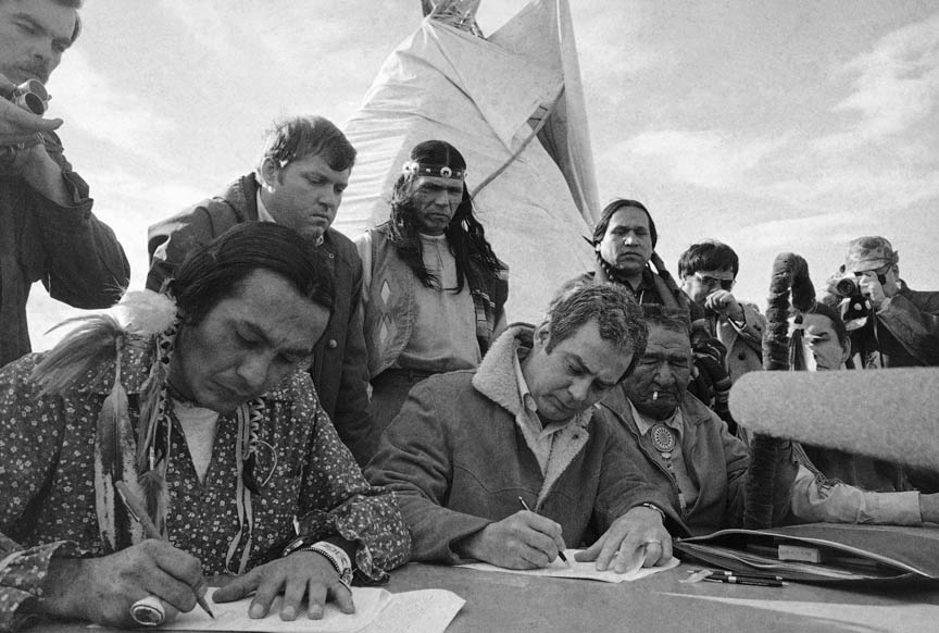 Russell Means, left, and assistant U.S. attorney general Kent Frizzell sign the Wounded Knee settlement on April 5, 1973 in South Dakota. Looking on left is Frizzell's assistant Richard Helstern and AIM leader Dennis Banks. 