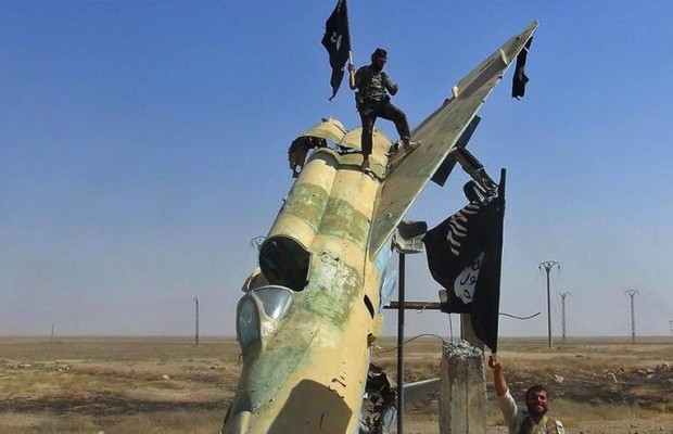 This undated image posted on Wednesday, Aug. 27, 2014 by the Raqqa Media Center of the Islamic State group, a Syrian opposition group, which has been verified and is consistent with other AP reporting, shows fighters of the Islamic State waving the group's flag from a damaged display of a government fighter jet following the battle for the Tabqa air base, in Raqqa, Syria. (AP Photo/ Raqqa Media Center of the Islamic State group)