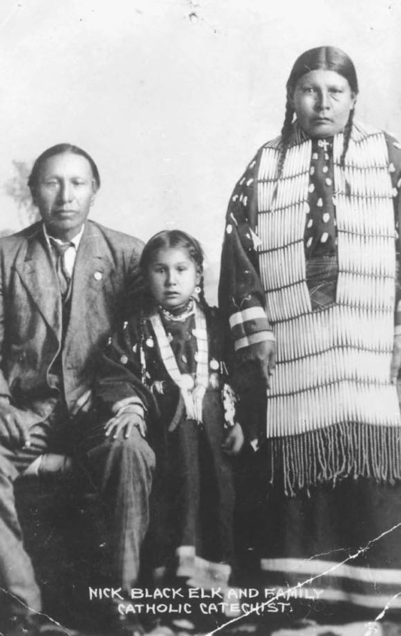 Nick Black Elk, daughter Lucy Black Elk and wife Anna Brings White, photographed in their home in Manderson, South Dakota, ca 1910. Black Elk wears a suit, his wife wears a long dress decorated with elk's teeth and a hair pipe necklace. Wikipedia